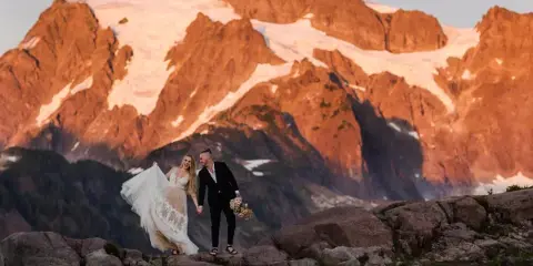 A bride and groom standing on a rocky mountain peak with a large snow-capped mountain in the background.