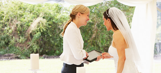 A wedding coordinator in a white shirt smiling and interacting with a bride in a wedding dress and veil.