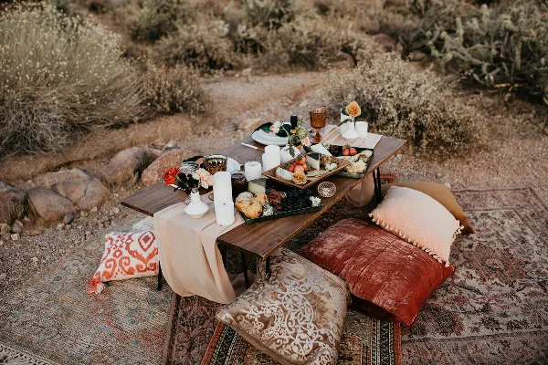 A styled outdoor event table with floral centerpieces, candles, and a food spread set on a rug with decorative pillows.