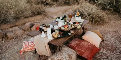 A styled outdoor event table with floral centerpieces, candles, and a food spread set on a rug with decorative pillows.