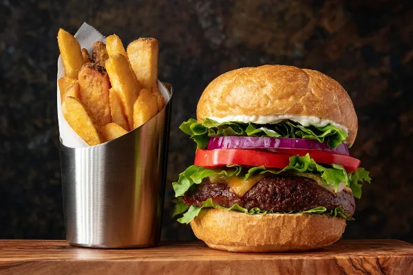 A cheeseburger with lettuce, tomato, and onion served next to a metal cup of french fries.