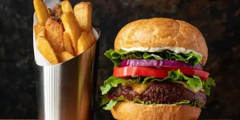 A cheeseburger with lettuce, tomato, and onion served next to a metal cup of french fries.