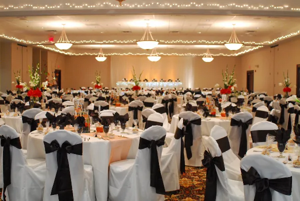 A wide shot of a large banquet hall set up for a wedding reception with round tables, white chair covers with black sashes, and floral centerpieces.