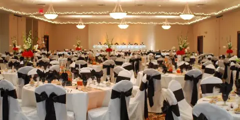 A wide shot of a large banquet hall set up for a wedding reception with round tables, white chair covers with black sashes, and floral centerpieces.