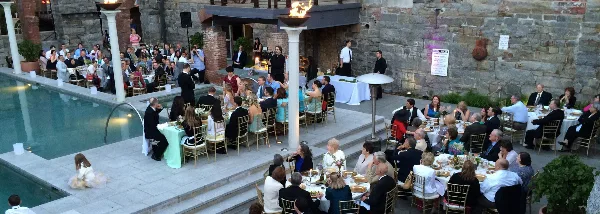 A large outdoor wedding reception with numerous guests seated at tables near a pool and stone wall.