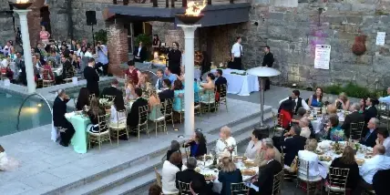A large outdoor wedding reception with numerous guests seated at tables near a pool and stone wall.