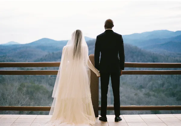 A bride and groom standing on a balcony overlooking a mountain range.