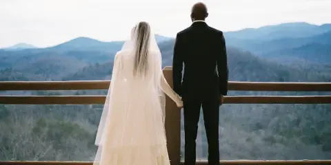 A bride and groom standing on a balcony overlooking a mountain range.