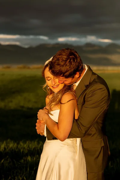 A bride and groom embracing in a field with mountains in the background.
