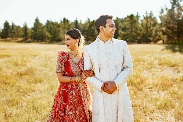 A bride in a red embroidered lehenga and a groom in a white sherwani standing in a grassy field.
