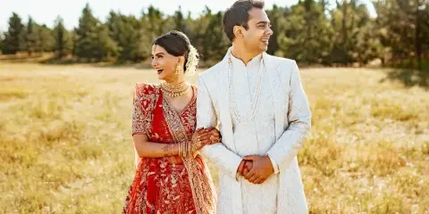 A bride in a red embroidered lehenga and a groom in a white sherwani standing in a grassy field.