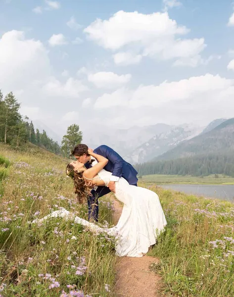 A bride and groom sharing a dip kiss in a wildflower meadow with mountains in the background.