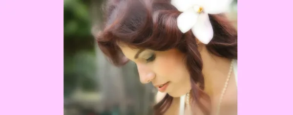 A bride with a white plumeria flower in her hair, looking down with a soft expression.