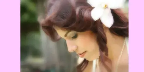 A bride with a white plumeria flower in her hair, looking down with a soft expression.
