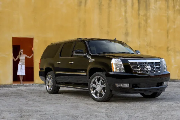 A black Cadillac Escalade ESV luxury SUV parked in front of a yellow stucco wall with a woman standing in a doorway.