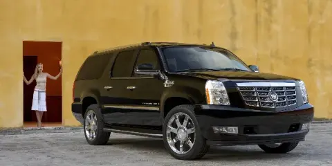A black Cadillac Escalade ESV luxury SUV parked in front of a yellow stucco wall with a woman standing in a doorway.