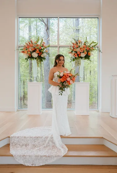 A bride in a white lace gown holds a bouquet of orange and white flowers while standing between two matching floral arrangements on pedestals in a bright room.