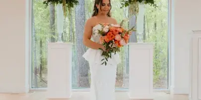 A bride in a white lace gown holds a bouquet of orange and white flowers while standing between two matching floral arrangements on pedestals in a bright room.
