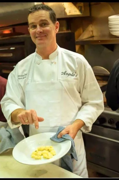Chef in a white coat embroidered with Angelo's holding a plated dish and a blue towel in a commercial kitchen.