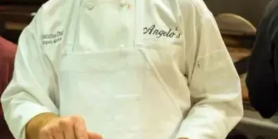 Chef in a white coat embroidered with Angelo's holding a plated dish and a blue towel in a commercial kitchen.