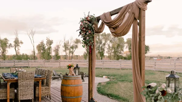 Outdoor wedding reception setup featuring a wooden arch with beige draping, floral arrangements, and a dining table with wicker chairs.