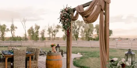 Outdoor wedding reception setup featuring a wooden arch with beige draping, floral arrangements, and a dining table with wicker chairs.