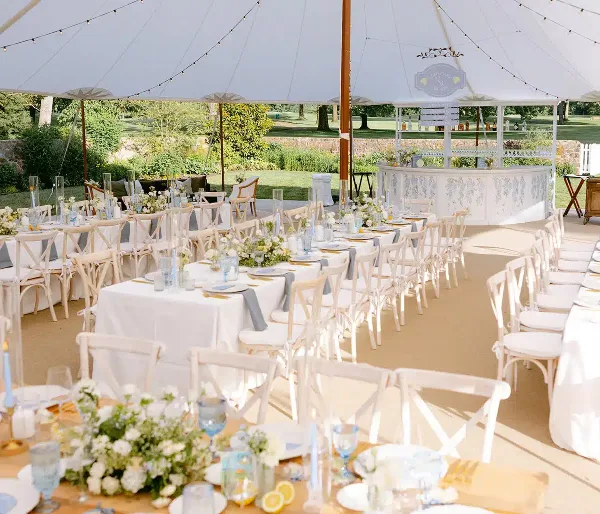 A wide shot of a white event tent set up outdoors with rows of dining tables, chairs, and a bar.