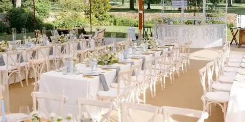 A wide shot of a white event tent set up outdoors with rows of dining tables, chairs, and a bar.