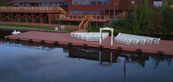 A wedding ceremony setup with white chairs and an arch on a wooden dock overlooking a lake, with a resort building in the background.