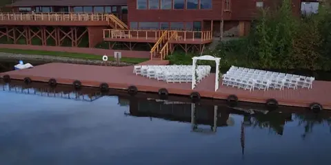 A wedding ceremony setup with white chairs and an arch on a wooden dock overlooking a lake, with a resort building in the background.