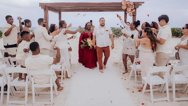 A bride and groom walking down a beach wedding aisle with guests and a decorated wooden arch.