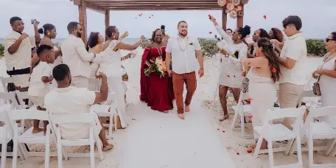 A bride and groom walking down a beach wedding aisle with guests and a decorated wooden arch.