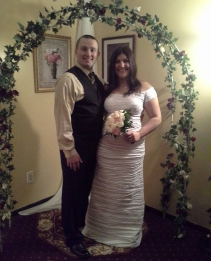 A bride and groom standing together under a floral arch in a wedding dress and suit.