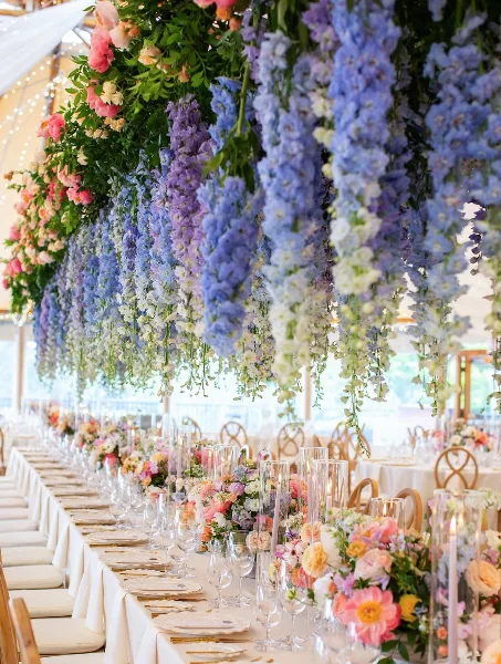 A long wedding reception table decorated with a massive overhead floral installation of blue and pink flowers and a table runner of mixed blooms.