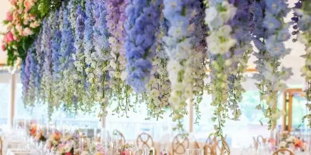A long wedding reception table decorated with a massive overhead floral installation of blue and pink flowers and a table runner of mixed blooms.