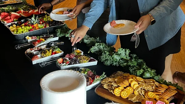 A catering staff member serving a plate of food from a buffet table featuring a charcuterie board, fruit platters, and crackers.
