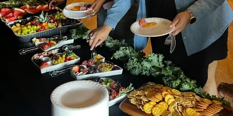 A catering staff member serving a plate of food from a buffet table featuring a charcuterie board, fruit platters, and crackers.