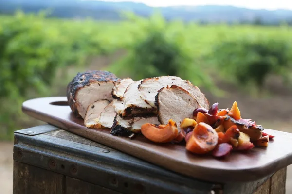 A plated dish of sliced roasted meat and roasted vegetables served on a wooden board in a vineyard setting.