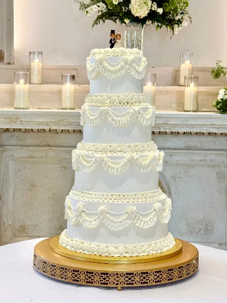 A four-tier white wedding cake decorated with elaborate piped swags and a bride and groom topper, displayed on a gold stand.