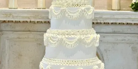 A four-tier white wedding cake decorated with elaborate piped swags and a bride and groom topper, displayed on a gold stand.