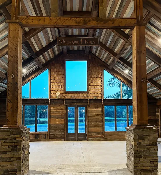 Interior view of the Whispering Pines rustic barn venue featuring a high wooden ceiling, stone fireplace, and large windows overlooking a pond.
