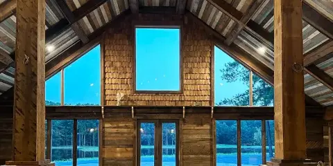 Interior view of the Whispering Pines rustic barn venue featuring a high wooden ceiling, stone fireplace, and large windows overlooking a pond.