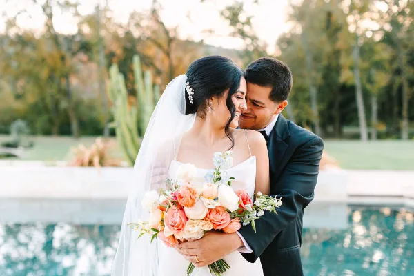 A bride and groom embracing outdoors near a pool, with the bride holding a bouquet of peach and white flowers.