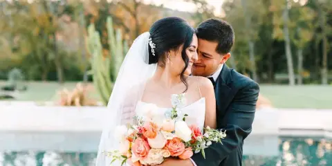 A bride and groom embracing outdoors near a pool, with the bride holding a bouquet of peach and white flowers.