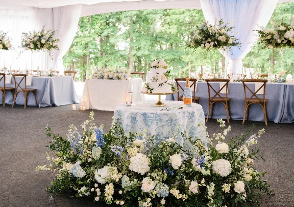 A wedding reception table setting featuring a floral centerpiece, a wedding cake, and blue table linens in a tented venue.