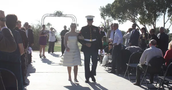 A bride and groom in a military dress uniform walk down the aisle at an outdoor wedding ceremony.