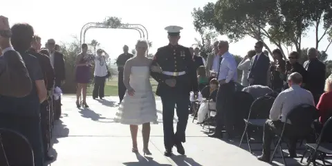 A bride and groom in a military dress uniform walk down the aisle at an outdoor wedding ceremony.