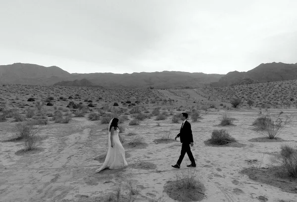 A black and white photograph of a bride and groom walking through a desert landscape with mountains in the background.