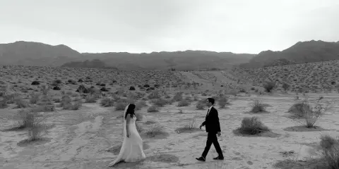 A black and white photograph of a bride and groom walking through a desert landscape with mountains in the background.