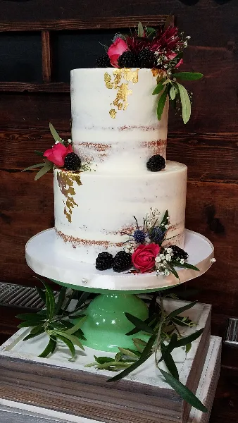 A two-tier semi-naked wedding cake with white frosting, gold leaf accents, and fresh floral and berry decorations on a green cake stand.
